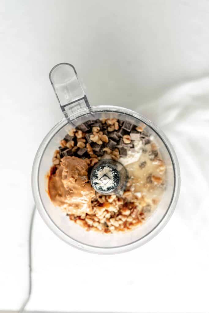 A food processor filled with walnuts, chocolate chunks, peanut butter, and other ingredients ready to be blended for the cookie dough mixture.