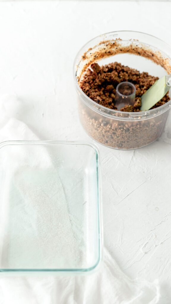 A food processor filled with crumbly cookie-dough mixture sits beside a glass dish that has been lightly greased with coconut oil.