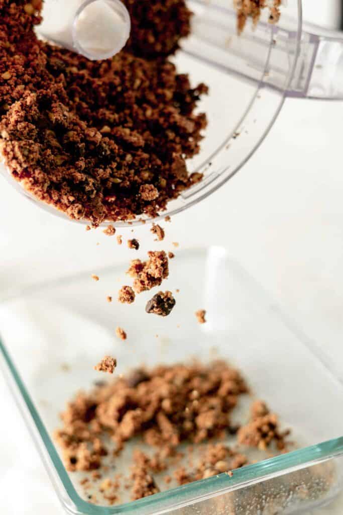 Crumbly cookie dough mixture being poured from a food processor into a glass baking dish.