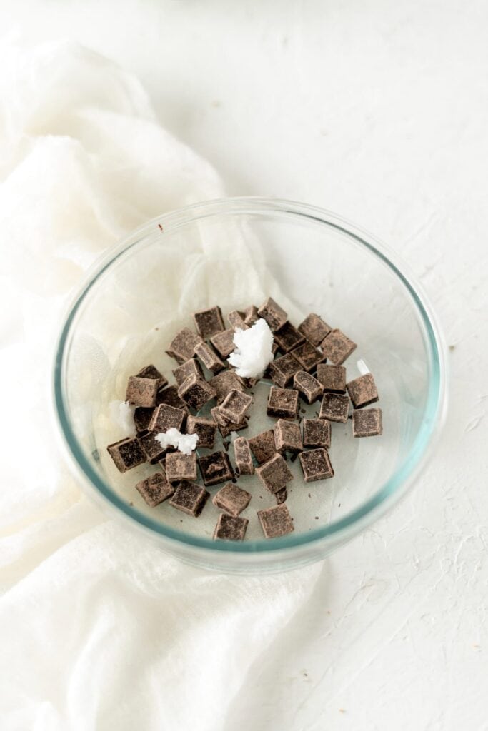 A glass bowl filled with chopped chocolate and small scoops of coconut oil, ready to be melted.