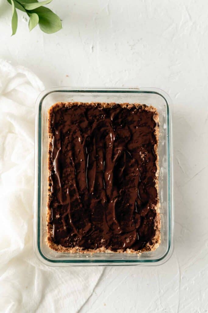 A thin layer of glossy melted chocolate is spread evenly over the pressed keto cookie-dough crust in a glass dish.