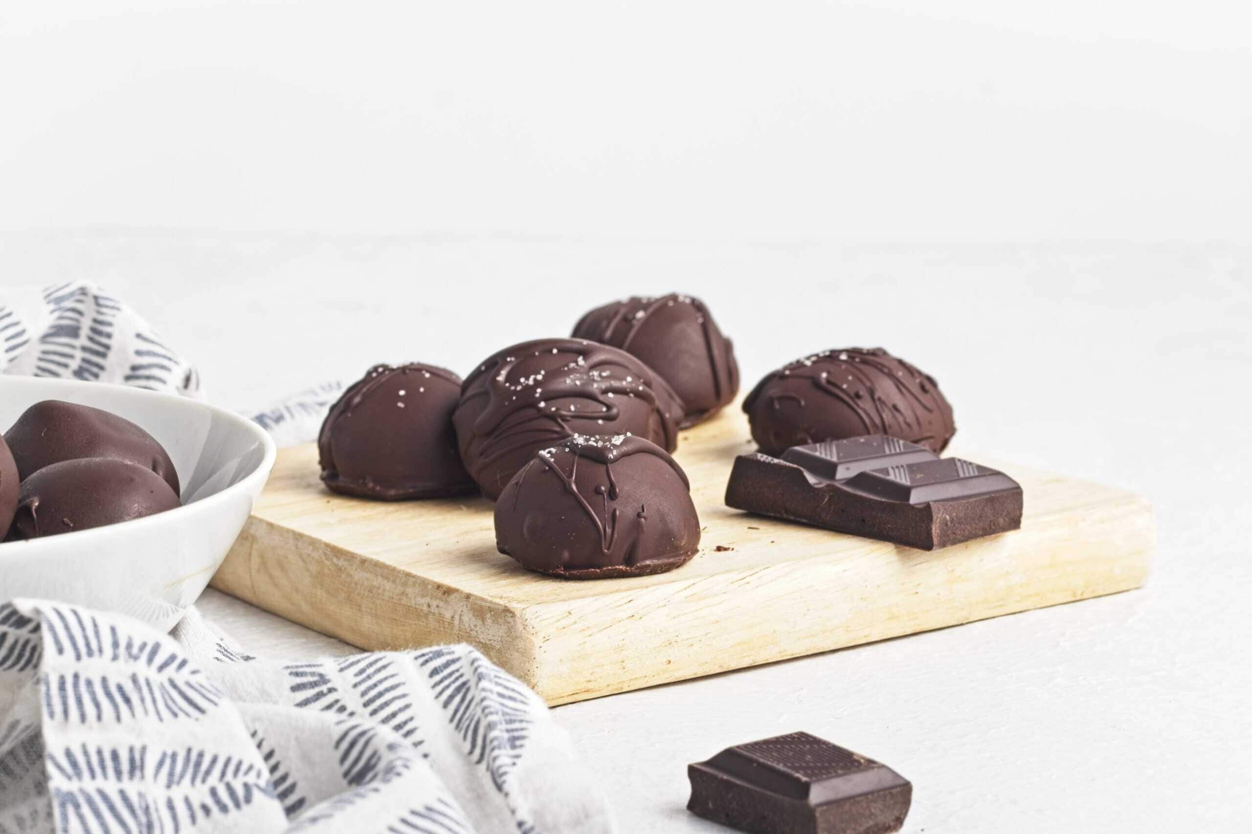 A small wooden board is topped with glossy chocolate-covered truffles, some drizzled and sprinkled with salt, alongside a few pieces of dark chocolate.