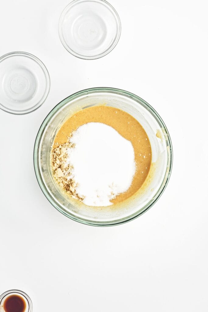glass mixing bowl with peanut butter mixture topped with a mound of protein powder, alongside small empty ingredient bowls on a white background.