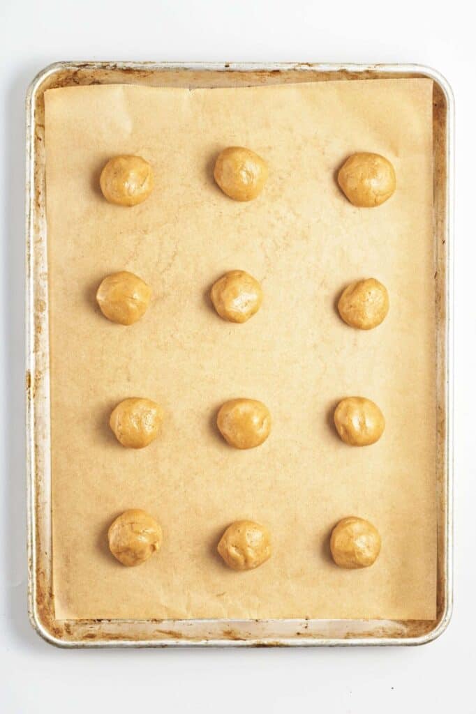 a parchment-lined baking tray holding evenly spaced round peanut butter cookie dough balls before flattening.
