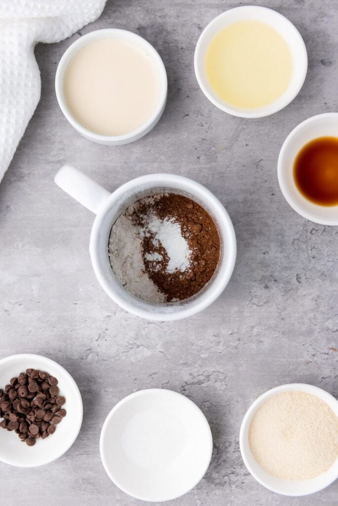 Ingredients for brownie-in-a-mug arranged around a mug filled with cocoa powder, flour, and sugar.