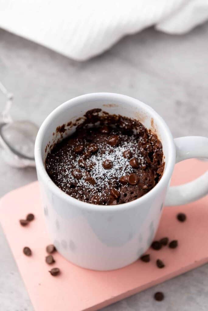 Chocolate mug cake topped with chocolate chips and a dusting of powdered sugar in a white mug.