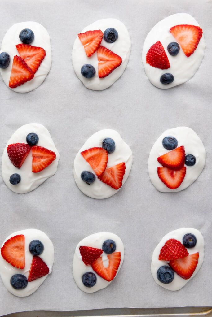Frozen yogurt bites on parchment paper topped with strawberries, and blueberries.