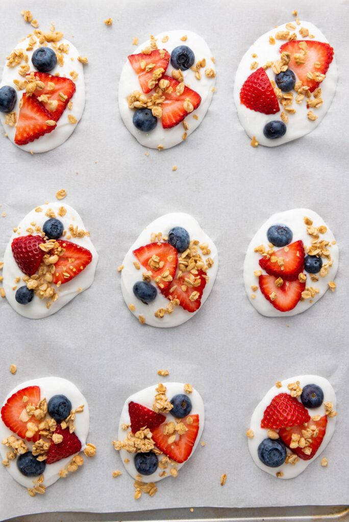 Frozen yogurt bites on parchment paper topped with granola, strawberries, and blueberries.
