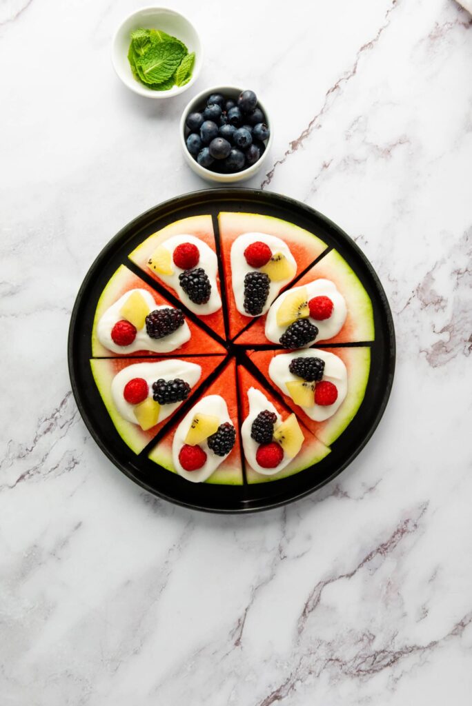 Watermelon pizza slices topped with yogurt, blackberries, raspberries, kiwi, and mint on a black plate with bowls of blueberries and mint nearby.