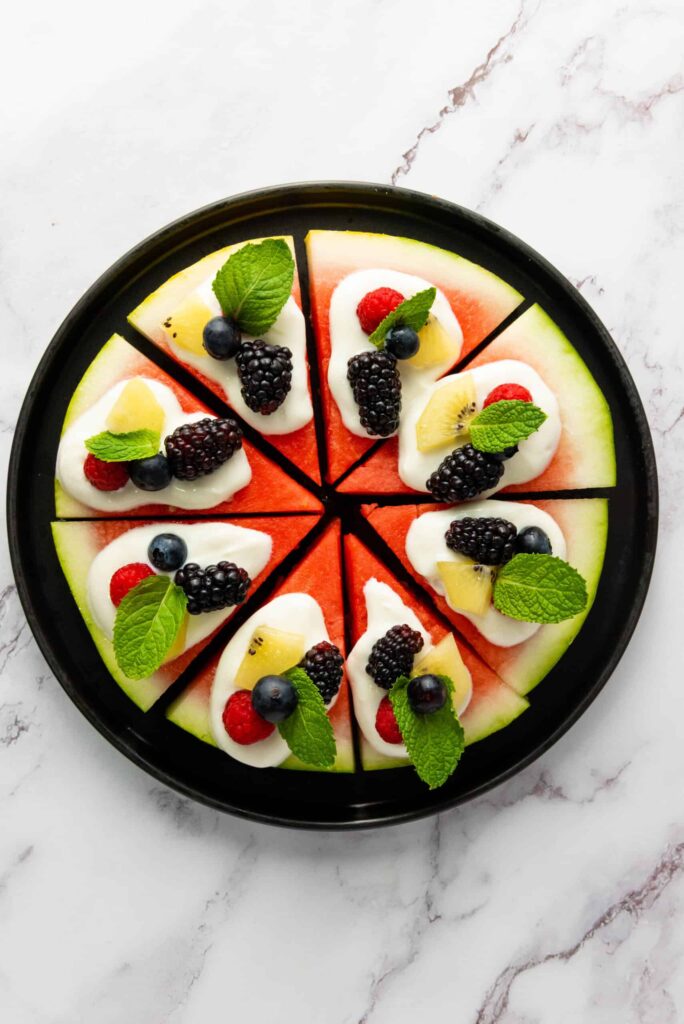 Overhead view of watermelon pizza decorated with yogurt, mixed berries, kiwi chunks, and fresh mint leaves.
