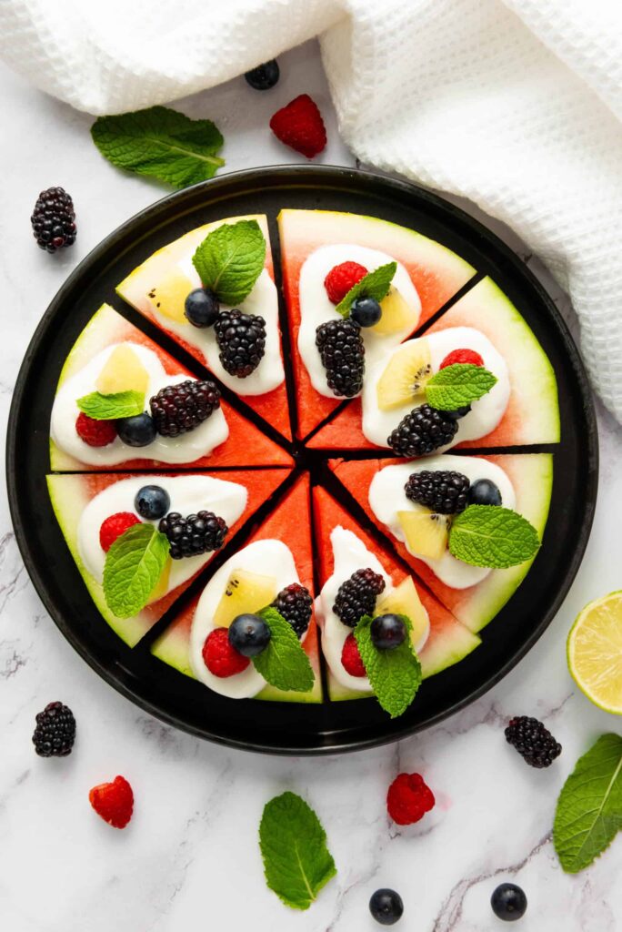 Watermelon pizza on a black plate surrounded by scattered berries, mint leaves, a lime half, and a white kitchen towel.