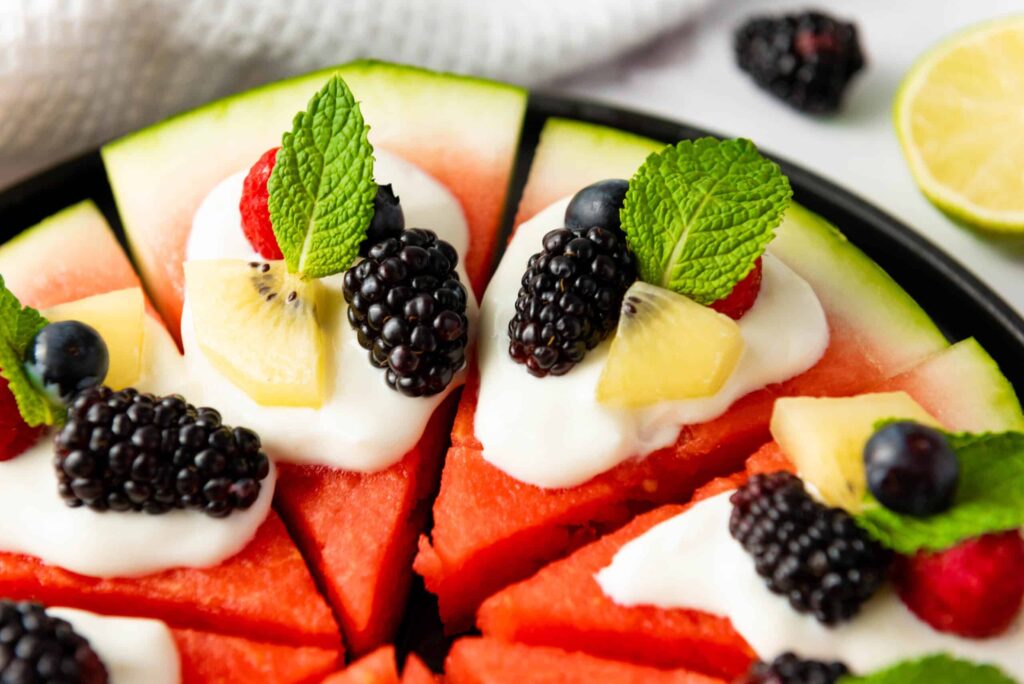 Close-up of watermelon pizza slices topped with yogurt, blackberries, blueberries, raspberries, kiwi, and mint.