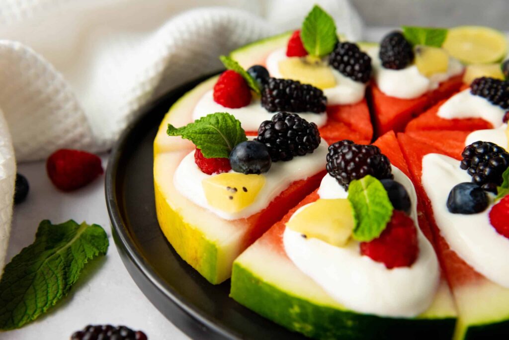 Watermelon pizza on a black plate surrounded by scattered berries, mint leaves, a lime half, and a white kitchen towel.