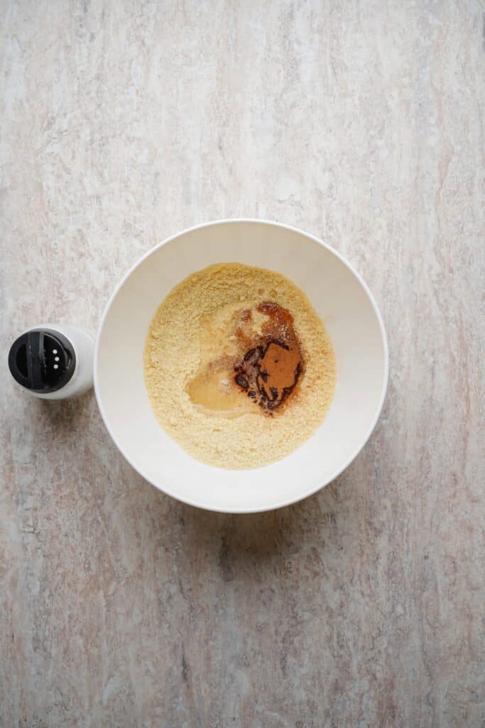 Bowl of almond flour and maple syrup with salt placed alongside on the surface.
