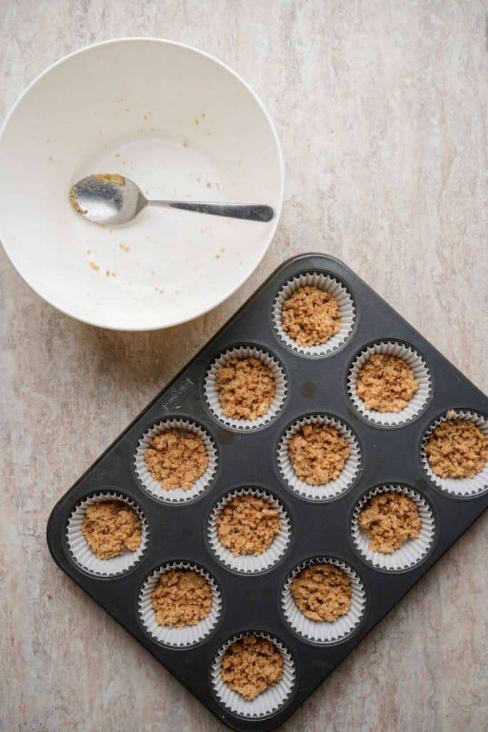 Almond flour crust mixture pressed into lined mini muffin cups in a baking tray, with an empty mixing bowl and spoon beside it.