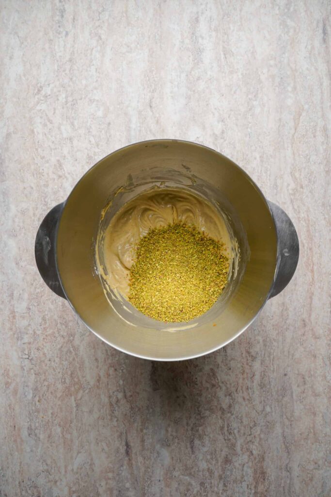Ground pistachios added to a creamy cheesecake mixture in a stainless steel mixing bowl.