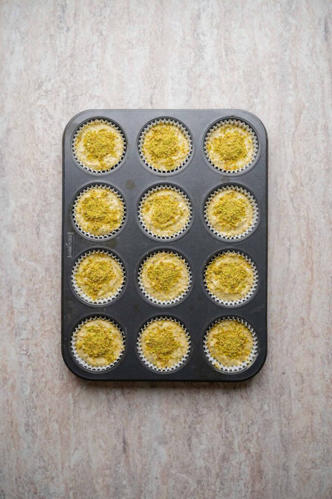 Unbaked mini cheesecakes topped with ground pistachios in a lined muffin tin, ready for the oven.