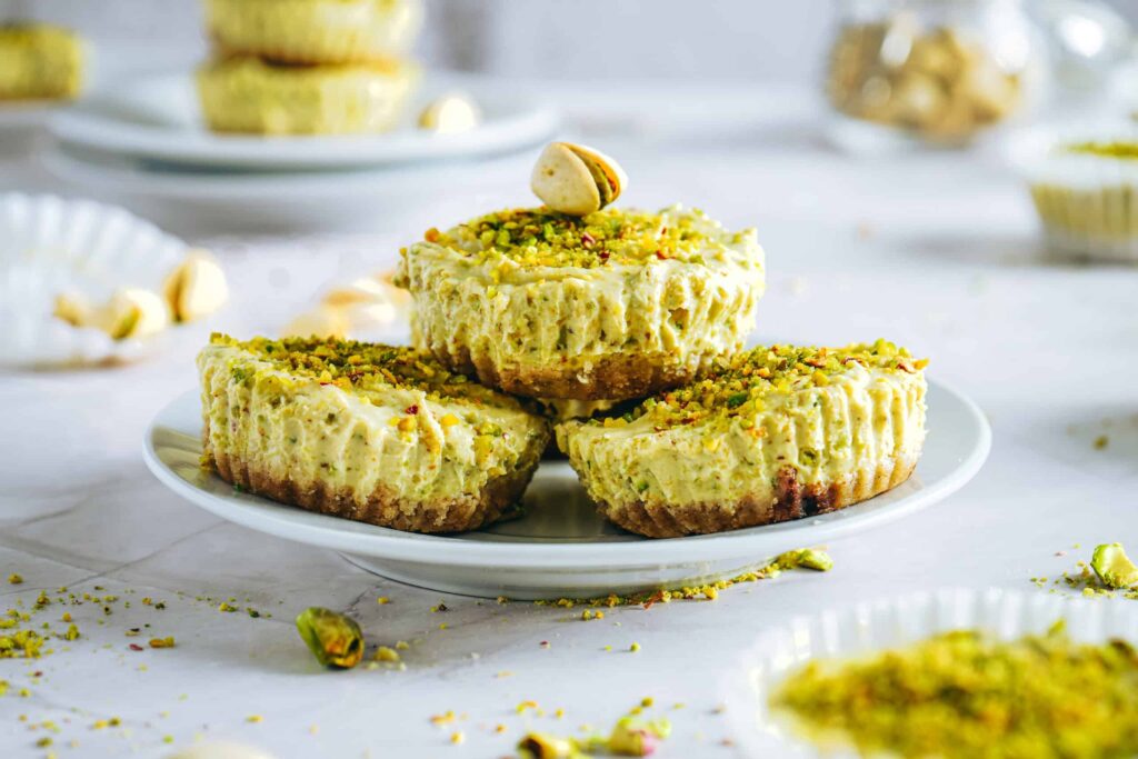 Stack of mini pistachio Greek yogurt cheesecakes topped with crushed pistachios on a white plate, showing a creamy texture and nutty crust.