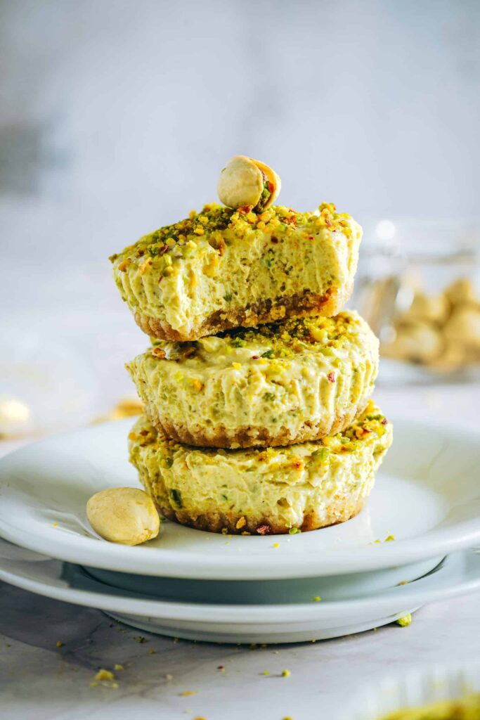 Stack of mini pistachio Greek yogurt cheesecakes topped with crushed pistachios on a white plate, showing a creamy texture and nutty crust.