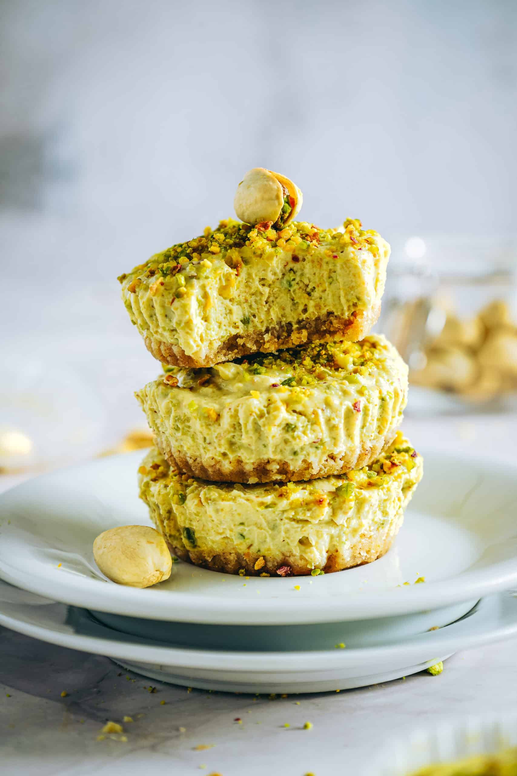 Stack of mini pistachio Greek yogurt cheesecakes topped with crushed pistachios on a white plate, showing a creamy texture and nutty crust.