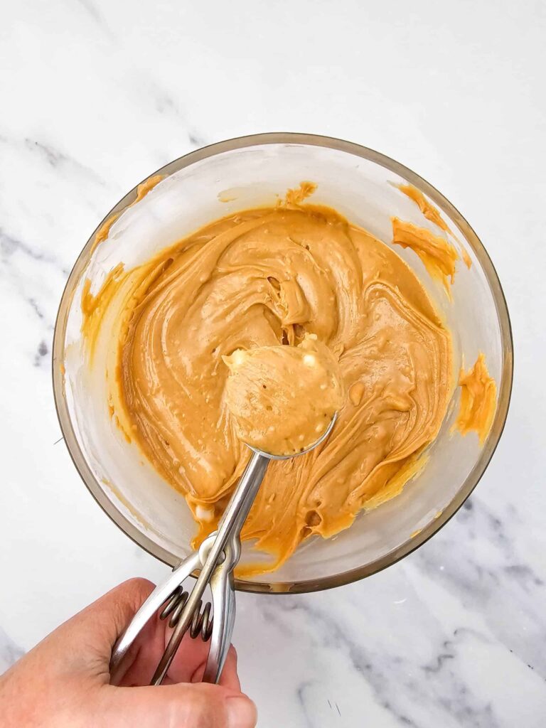 Peanut butter mixture being scooped with a small cookie scoop over a glass bowl.