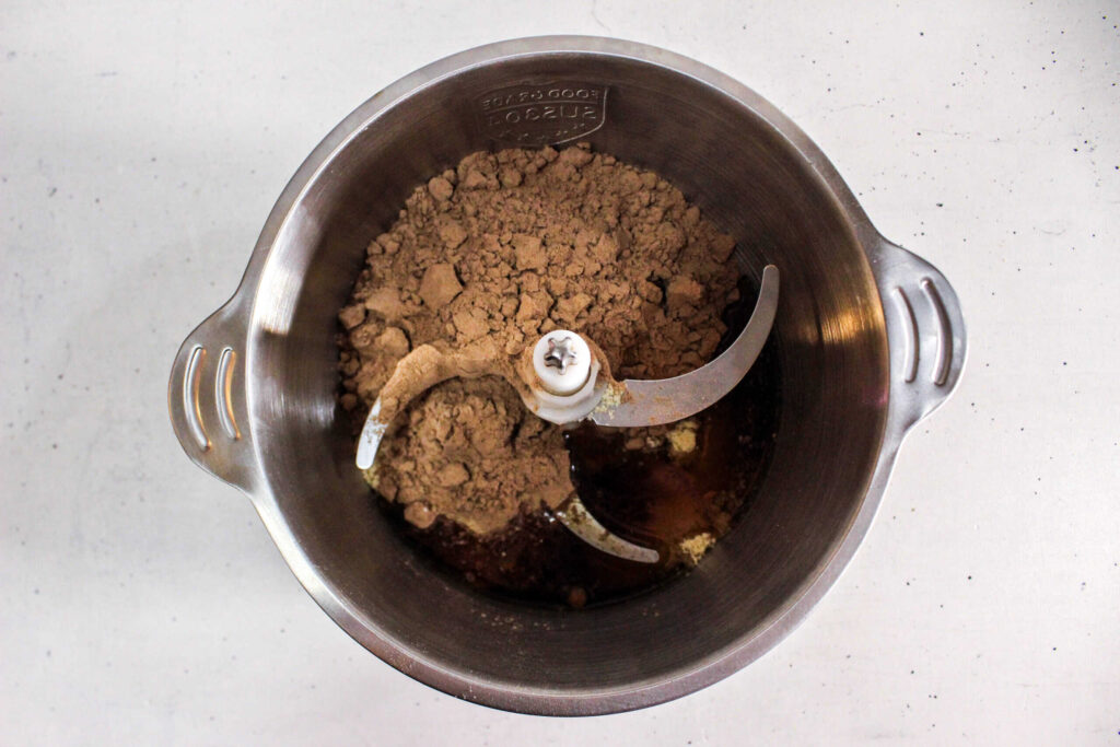 Chocolate protein powder mixture with maple syrup in a food processor bowl before blending