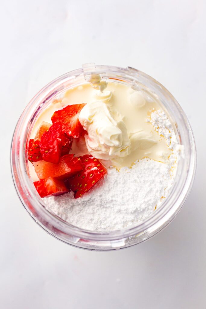 Overhead view of strawberry cheesecake ice cream mixture with cream, powdered sugar, and fresh strawberries in a blender cup.