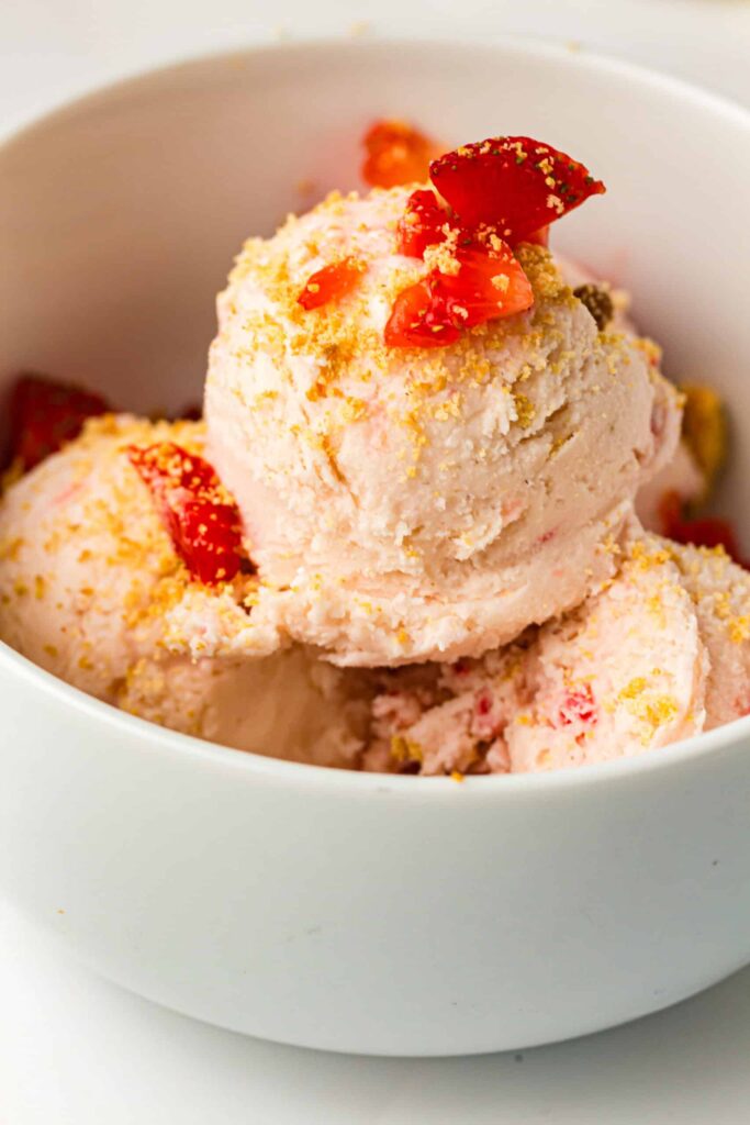 Soft strawberry ice cream scoops garnished with chopped strawberries and sprinkled biscuit crumbs in a white bowl.